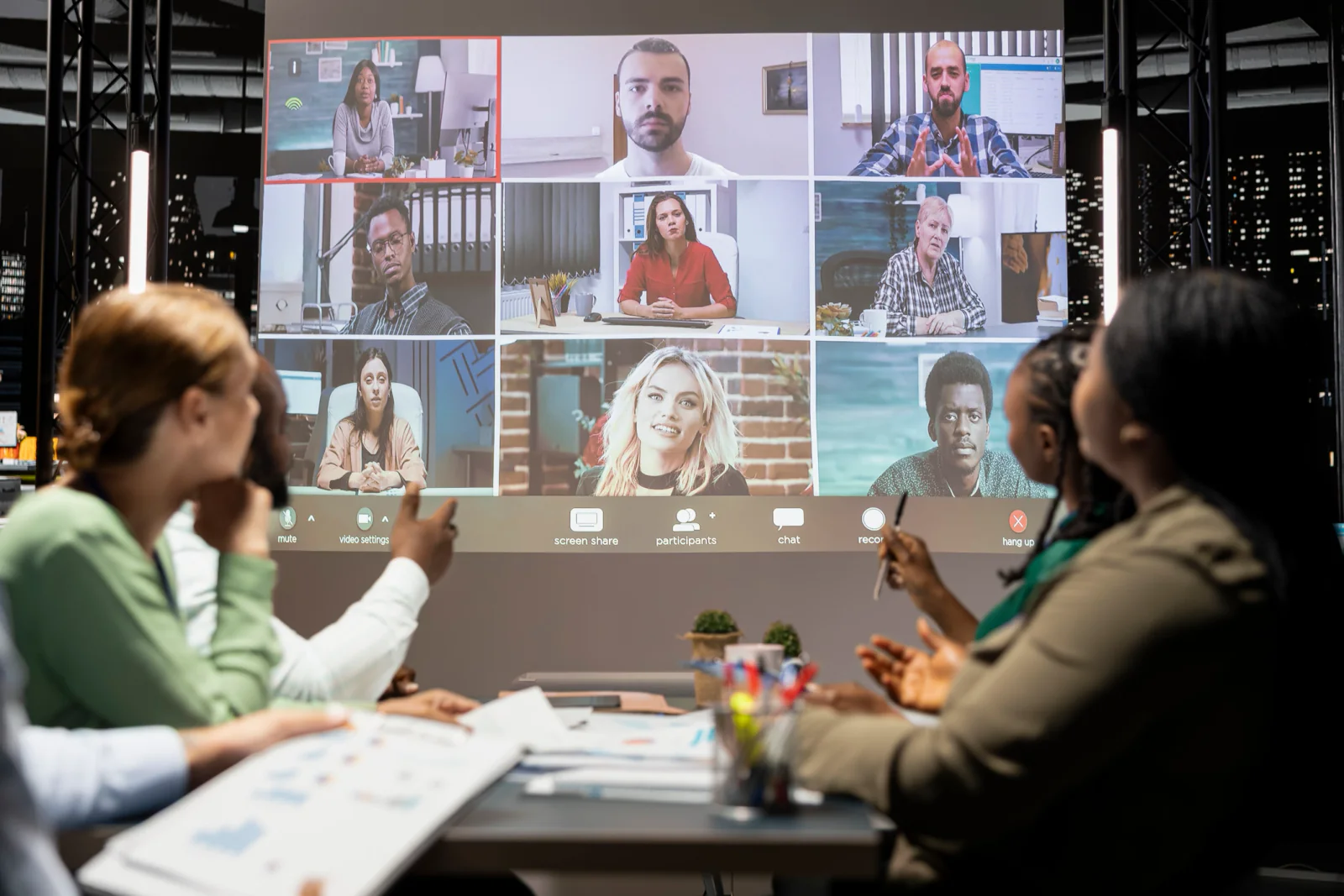 Hybrid meeting room with in-person attendees, large screens showing remote participants, cameras, microphones, and conferencing equipment