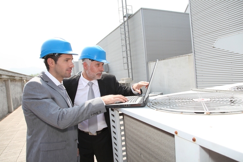 Two mean wearing hardhats point at a laptop screen while standing on the roof of a building