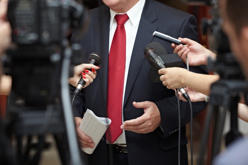 Several reports holding different microphones up to an individual wearing a suit with a red tie