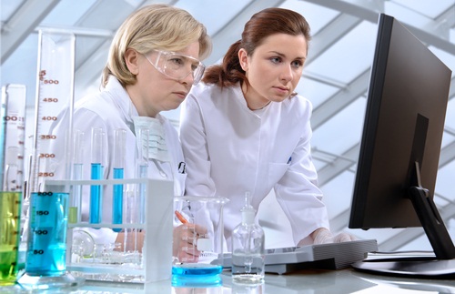 Two scientsists wearing labcoats look at a computer screen. Various chemicals in vials and beakers lay on the tbale next to them