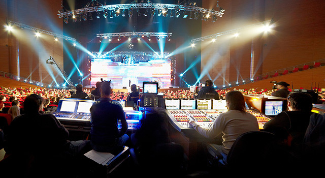 Sound technicians sitting at a mixing board while a large event occurs on stage in front of them