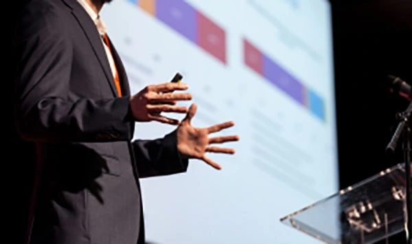 A man giving a presentation at a podium with a large projection screen displaying information behind him