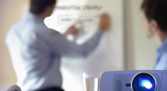 Individuals working on a whiteboard while a projector in the foreground is shown illuminating